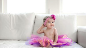 Cute Baby Girl Is Sitting On White Couch Wearing Colorful Skirt And Woolen Knitted Flower Cap HD Cute