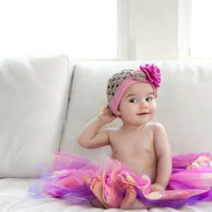 Cute Baby Girl Is Sitting On White Couch Wearing Colorful Skirt And Woolen Knitted Flower Cap HD Cute