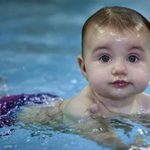 Cute Baby Is Swimming On Body Of Water Wearing Purple Shorts In A Blur Water Background HD Cute