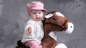 Cute Baby Is Sitting On Toy Horse Wearing White Top And Pink Pant And Cap In Ash Background HD Cute