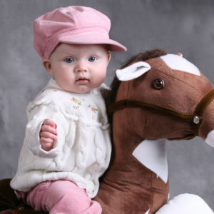 Cute Baby Is Sitting On Toy Horse Wearing White Top And Pink Pant And Cap In Ash Background HD Cute Wallpapers