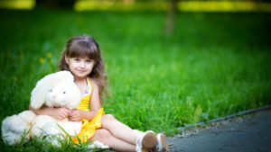 Cute Little Girl Is Wearing Yellow Dress Sitting On Green Grass In Blur Green Background With Teddy Toy HD Cute