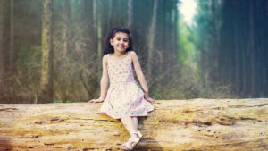 Smiley Cute Girl Is Sitting On Tree Trunk Wearing White Flowers Printed Dress In Forest Background HD Cute