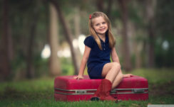 Smiley Cute Little Girl Is Wearing Blue Dress Sitting On Red Suitcase In Blur Trees Background HD Cute Wallpapers