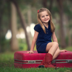 Smiley Cute Little Girl Is Wearing Blue Dress Sitting On Red Suitcase In Blur Trees Background HD Cute Wallpapers