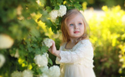 Little Cute Ash Eyes Girl Is Wearing White Netted Dress Standing Near Green Plant Touching White Flower HD Cute
