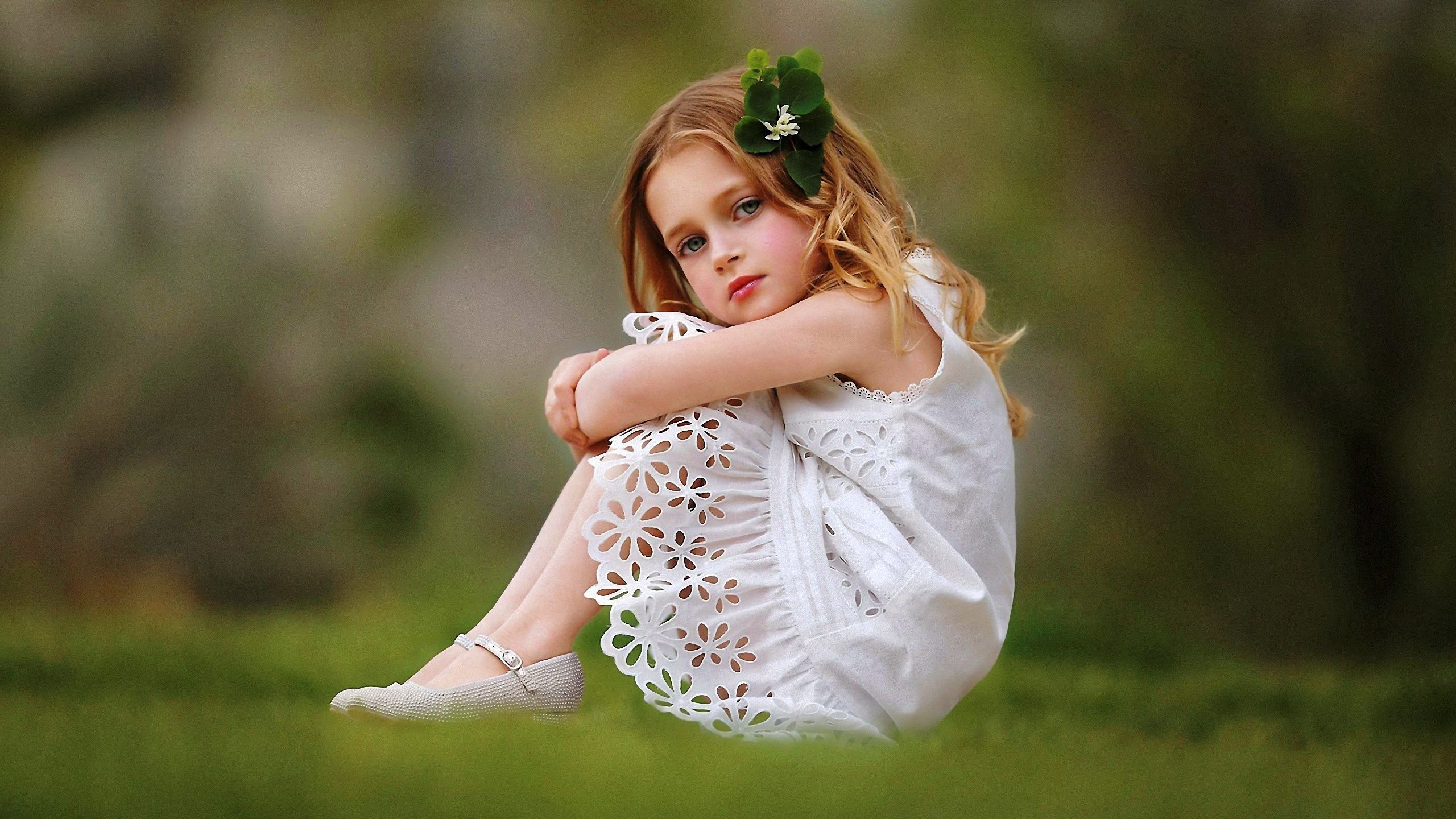 Cute Girl Baby Is Wearing White Dress Sitting On Grass Facing One Side And Having Green Leaves On Head In A Blur Background HD Cute