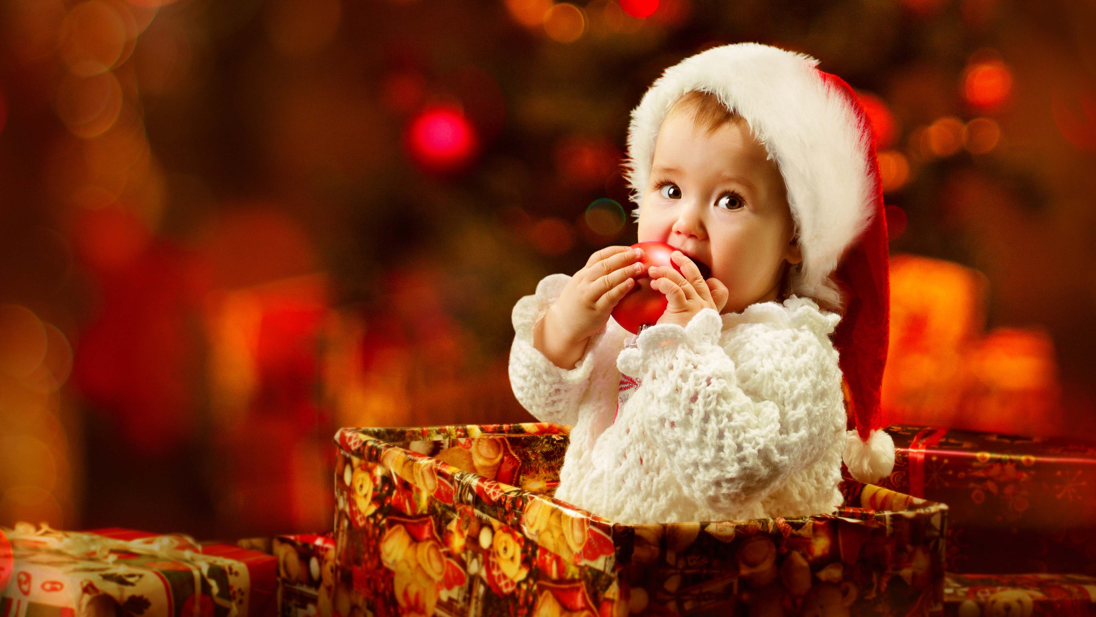 Cute Girl Baby Inside Gift Box Wearing Santa Cap And Sandal Color Dress Biting Apple In Blur Light Background 4K HD Cute